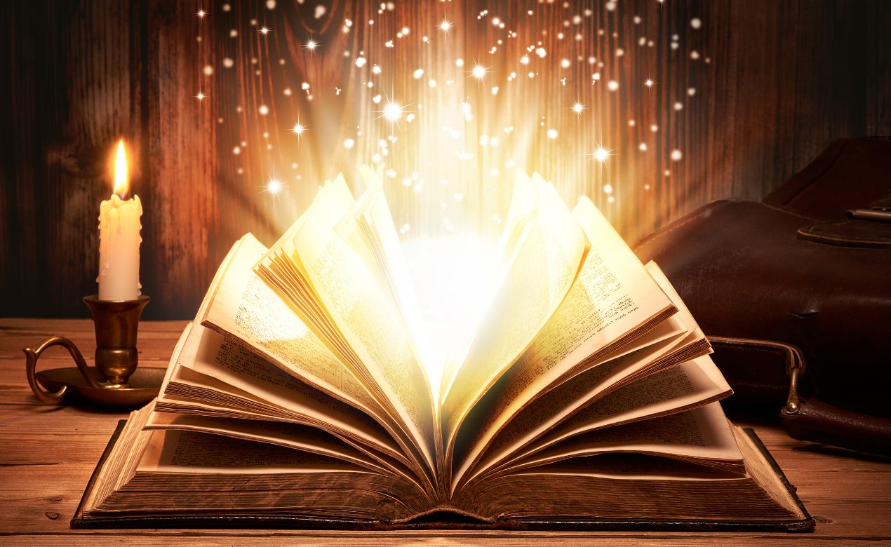 An open book with flashes and sparks of light emitting from it. The book is sitting on a wooden desk with a candle in a candleholder sitting beside it. The scene has a magical, fantastical feel to it.