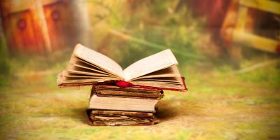 An open book sits on top of other books that are closed. The books sit on a grassy surface. Blurs of light swirl around the books. Blurs of indescribable red shapes make up the background.