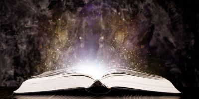 An open book sits on a table with light and purple and gold mist emitting from it.