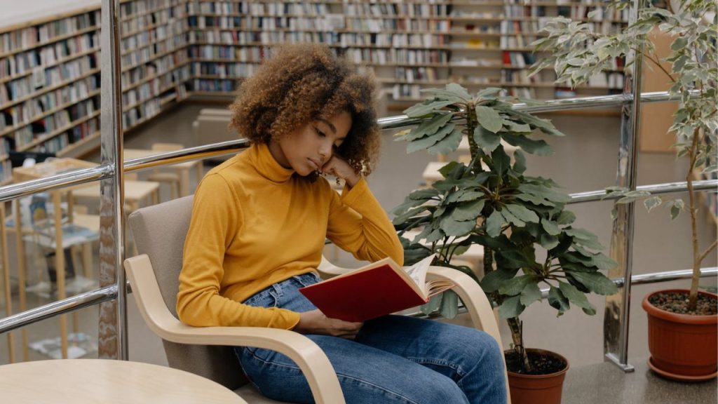 A black woman in mustard high neck tshirt and blue denims reads in a library.