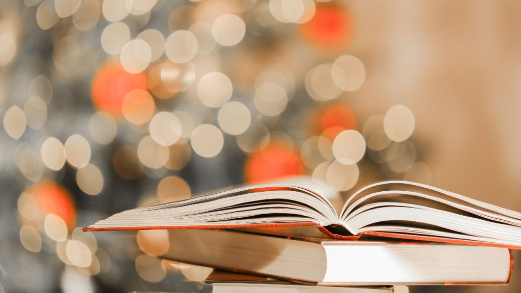 An open book in front of a blurred Christmas tree with red ornaments and white lights.