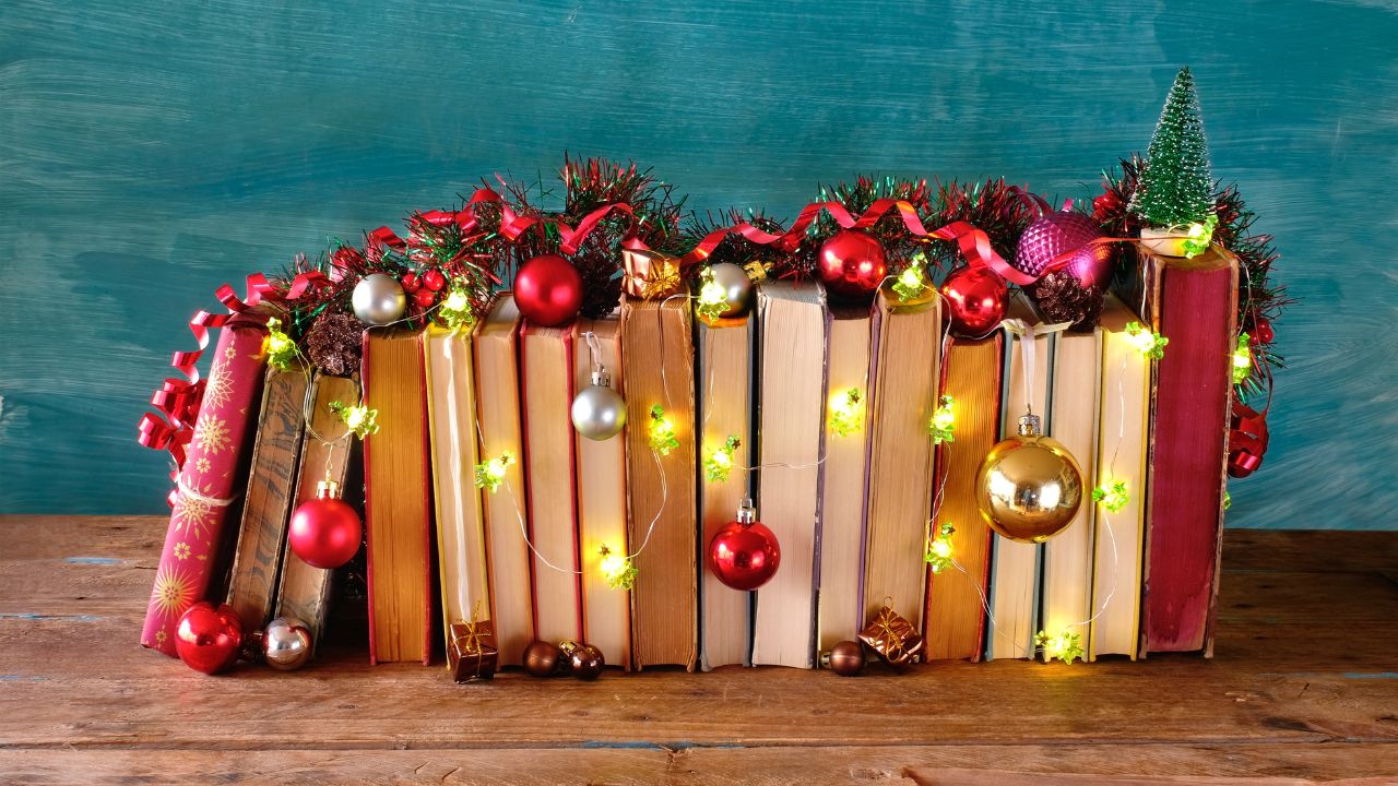 A row of books turned on their sides draped in multicolored Christmas lights and Christmas tree ornaments.
