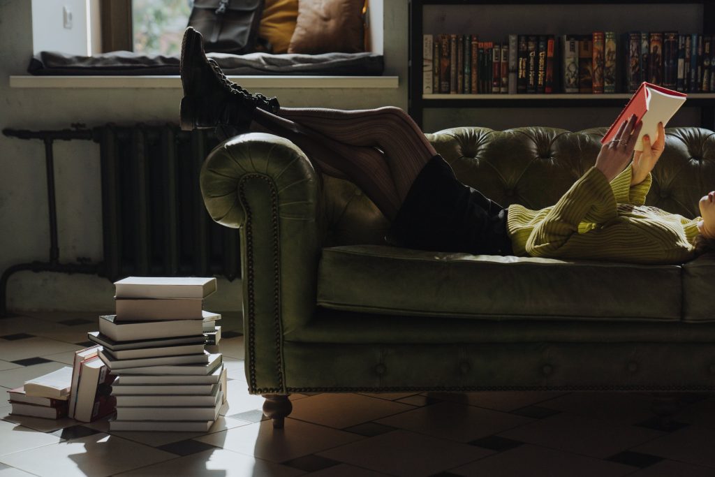 Person lounging on a sofa reading a book. A large stack of closed books at the end of the sofa.