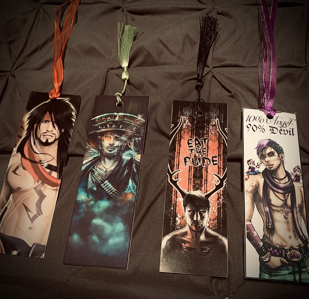 There are four bookmarks featuring males in various looks, colors, and clothing. Two of the four have words on them. They are set against a gray cloth surface.