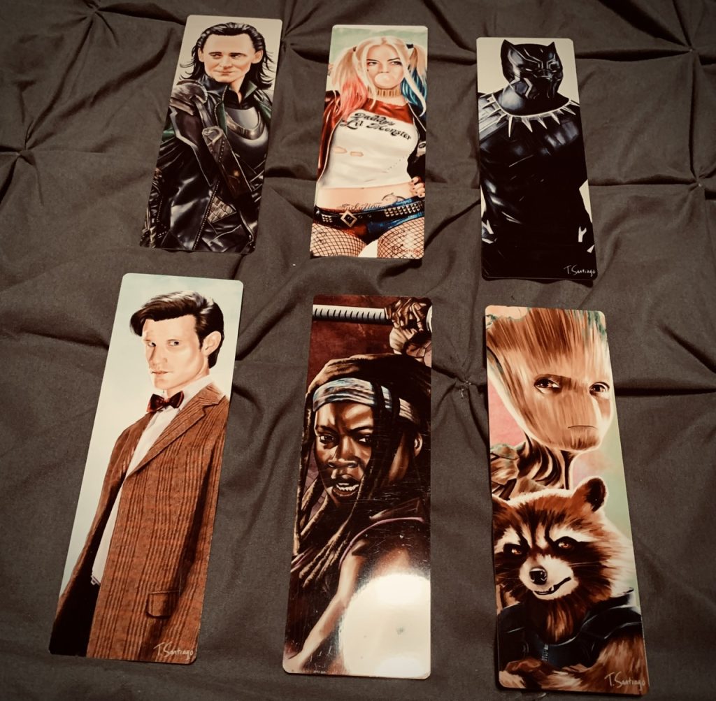There are six bookmarks featuring the liknesses of various popular tv, movie, and book characters - Loki, Harley Quinn, Black Panther, Doctor Who, Michone, And Groot and Rocket. They are set against a gray cloth surface.