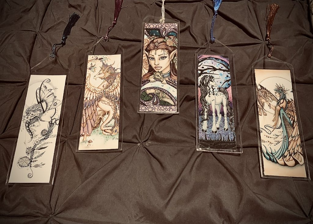 Five bookmarks with various depictions of fairies, dragons, and unicorns. Four of the five are colorful bookmarks. The fifth one is black and white. They are set against a gray cloth surface.