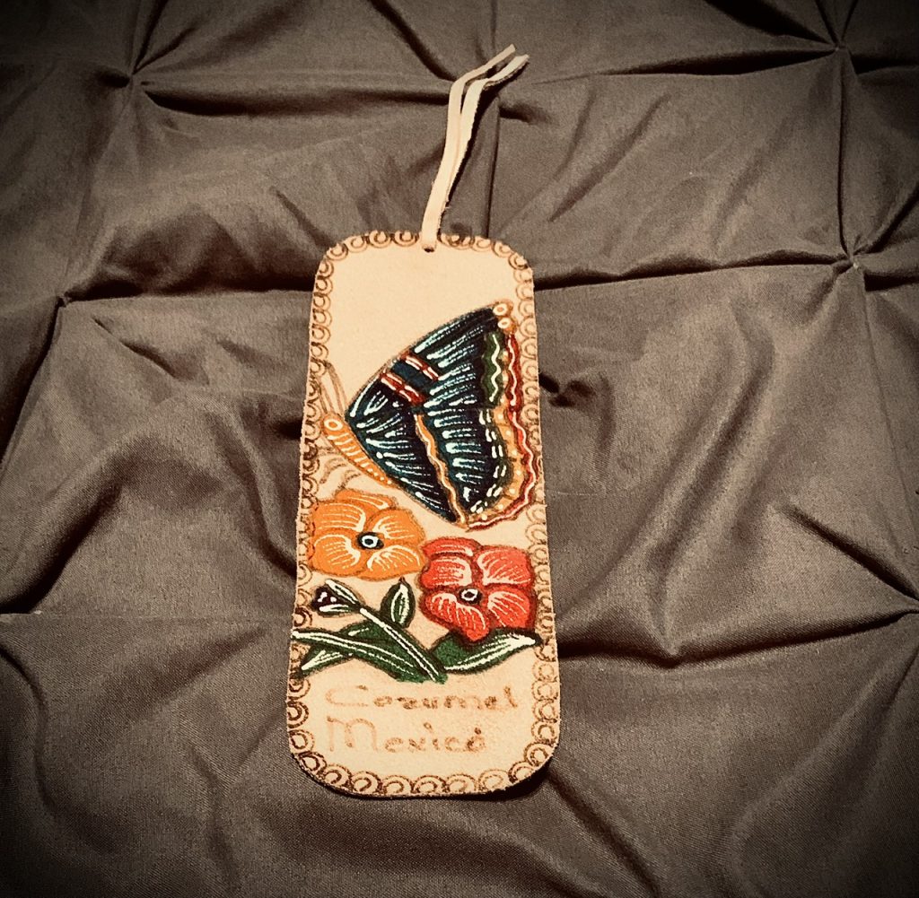 There is a large beige cloth bookmark with a butterfly and two flowers stenciled on it, and swirls are drawn around the border of the bookmark. "Cozumel, Mexico" is written under the  drawn image. The bookmark is set against a gray cloth surface.