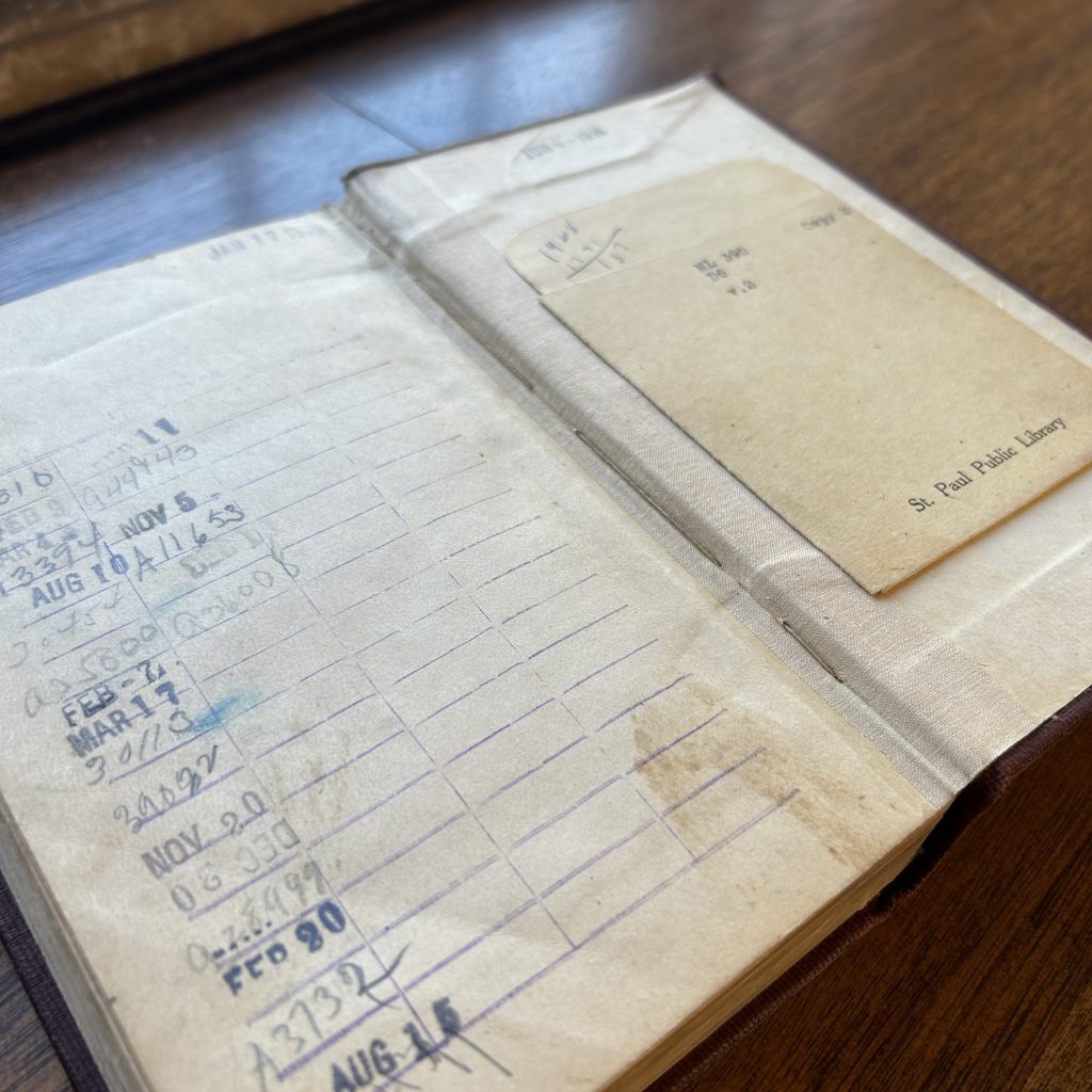 An open book with dates stamped on lines running down the page on the left side. On the right side, a library card pocket with words and numbers on it. The book sits on wooden table.