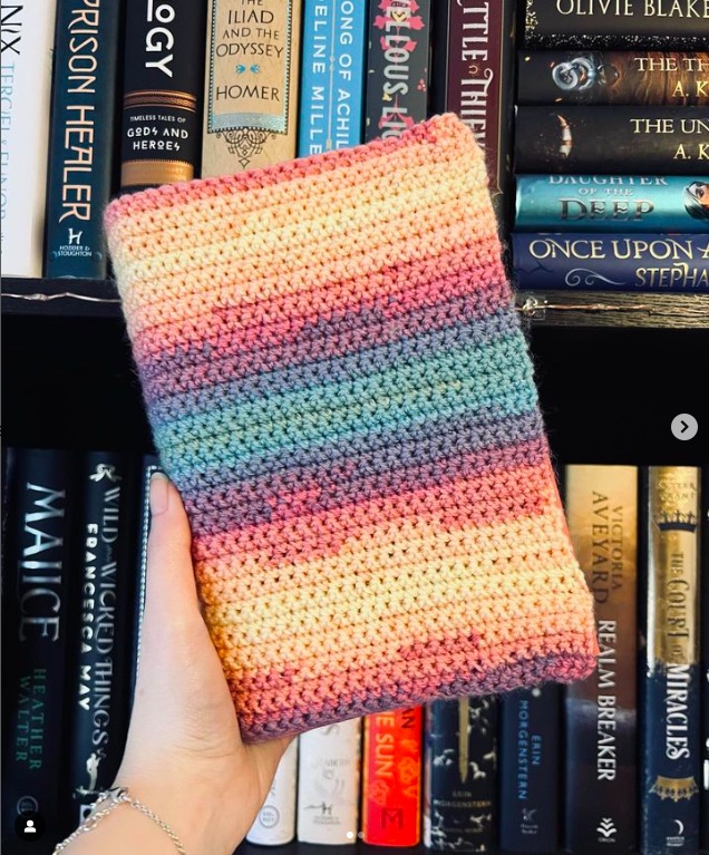A hand holding up a book in a knitted book cover in front of a full bookshelf.
