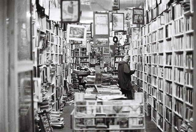 A black and white bookstore with books on shelves and stacked in piles. There is a man in a sweatshirt standing and reading a book.