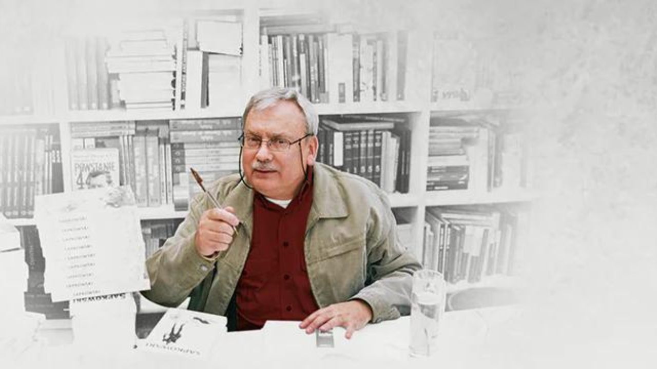 Andrzej Sapkowski, author of The Witcher, sitting in front of a white and gray bookshelf. He is wearing a redish-brown shirt and a beige jacket.