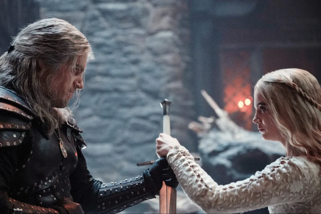 Henry Cavill as Geralt and Freya Allan as Ciri holding a sword. Geralt is wearing black and Ciri is wearing white.