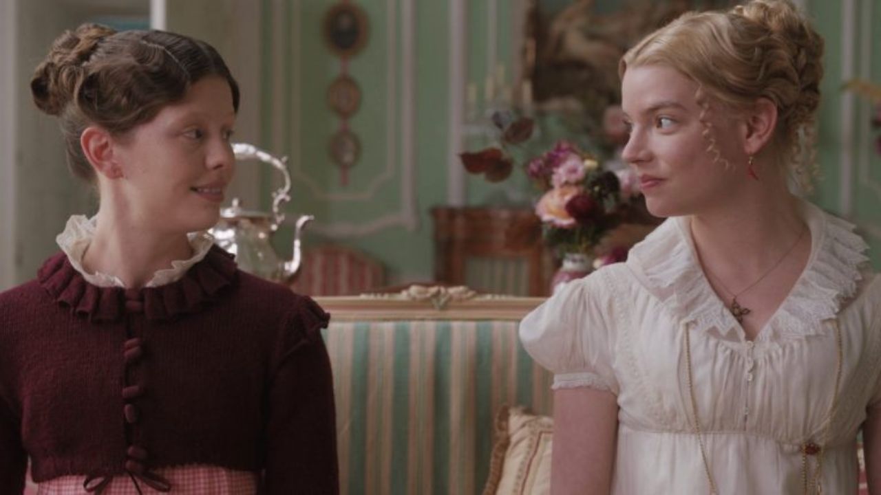 Emma and Harriet from the 2020 film 'Emma.' sitting in a colorful room and smiling while looking at each other.