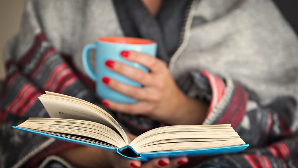 A person wrapped in a blanket holding a book and a mug.