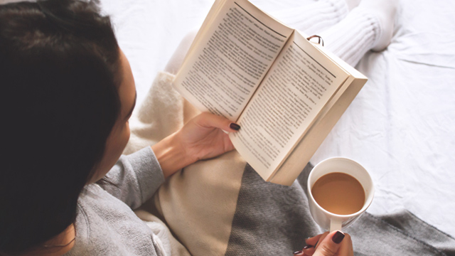 woman reading while holding up cup of coffee while laying in bed