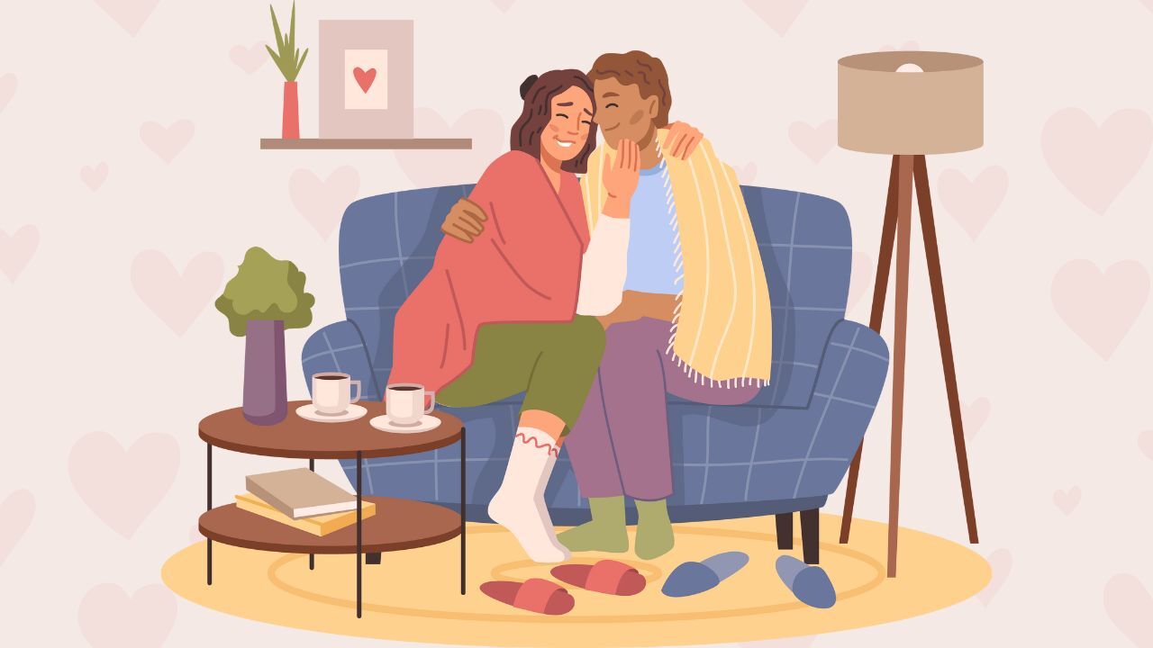 A couple sits on a blue couch, snuggling close. They have their arms wrapped around each other in a warm embrace. They are sitting a living room setting, with a coffee table in front of them and two cups of liquid. Their slippers are off, and behind them is a white wall with a vase and picture on a shelf.
