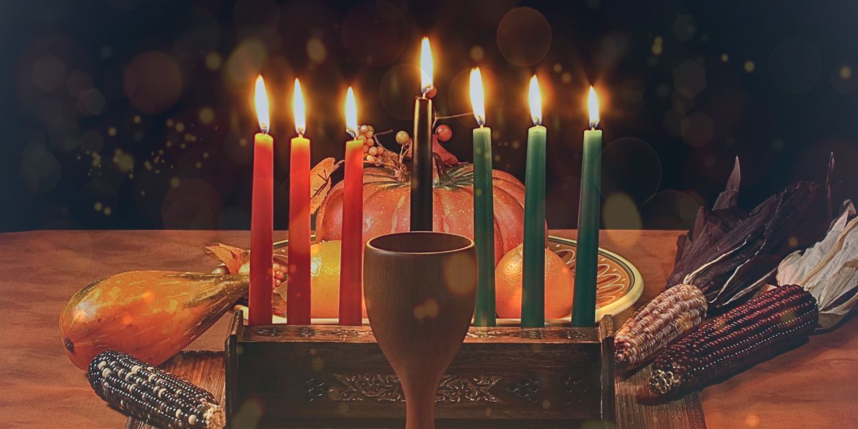 Seven candles in a candleholder are lit up. a cup sits in front of the candleholder. Food sits behind the candleholder. The background is a calm black color. Everything is set up on a wooden table.