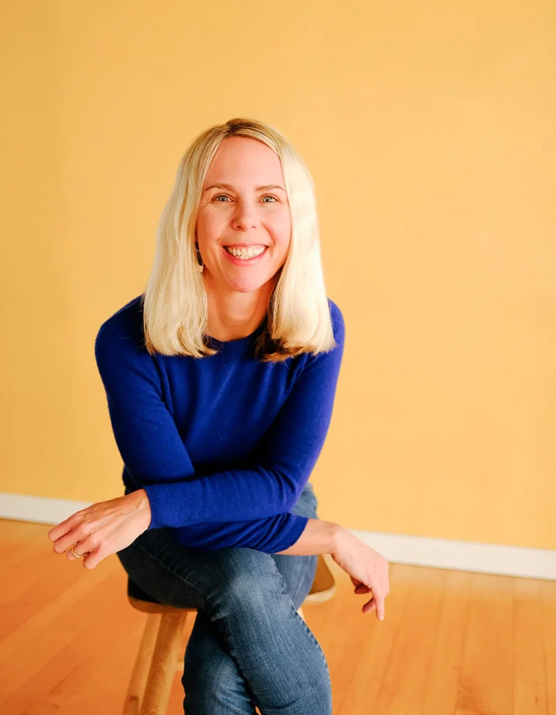 Polly Stewart sitting in front of a yellow wall and smiling.