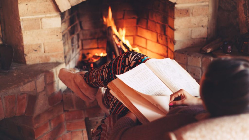 Image of a person sitting next to a warm fire with a book in hand. 