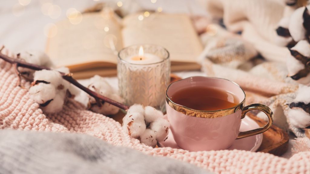 Image of a candle, tea and a book sitting next to some flowers and a blanket. 
