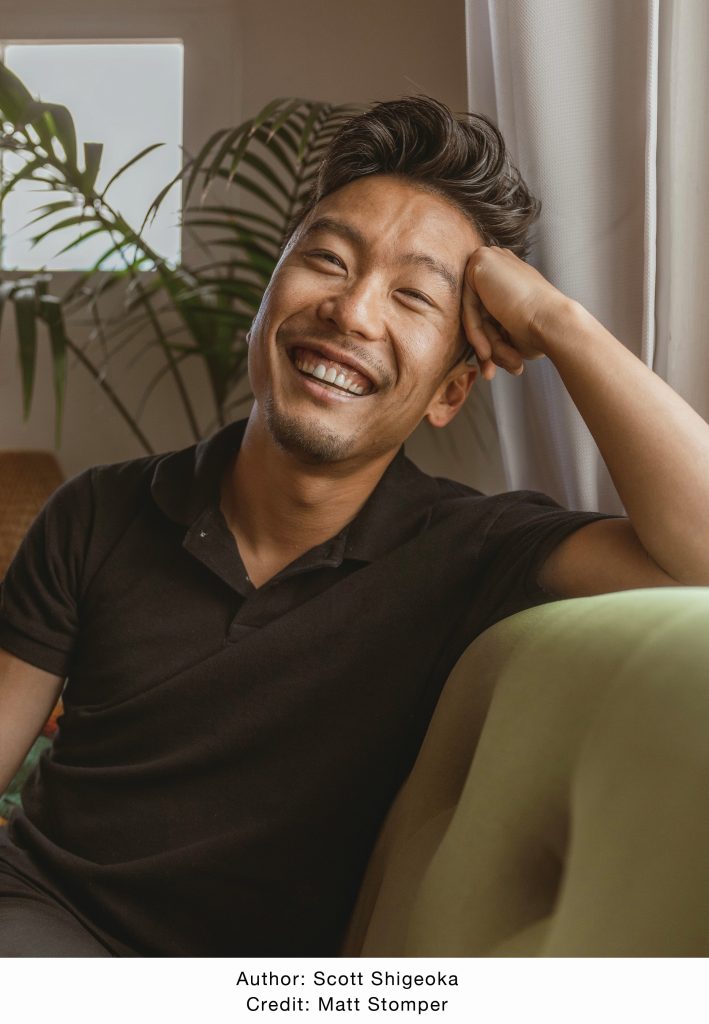 Scott Shigeoka leaning against a green couch and smiling widely.