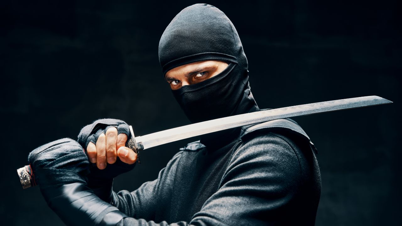 A closeup of a ninja almost completely covered in black clothing and holding a sword over his shoulder