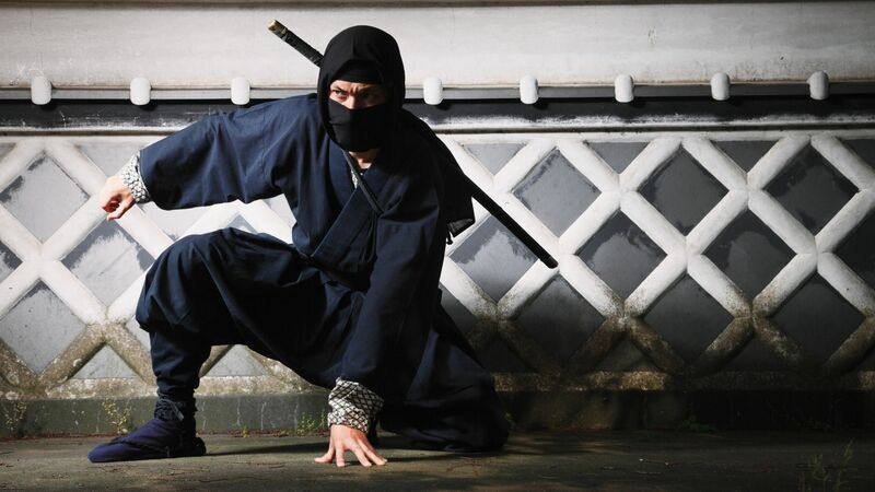 A ninja wearing all black kneeling with a sword strapped to his back