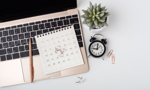 A rose gold laptop on a white table also with a calendar, clock, pen and plant