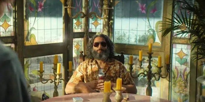 A man with gray hair and a beard wearing sunglasses and drinking a diet coke as he is surrounded by yellow candles. 