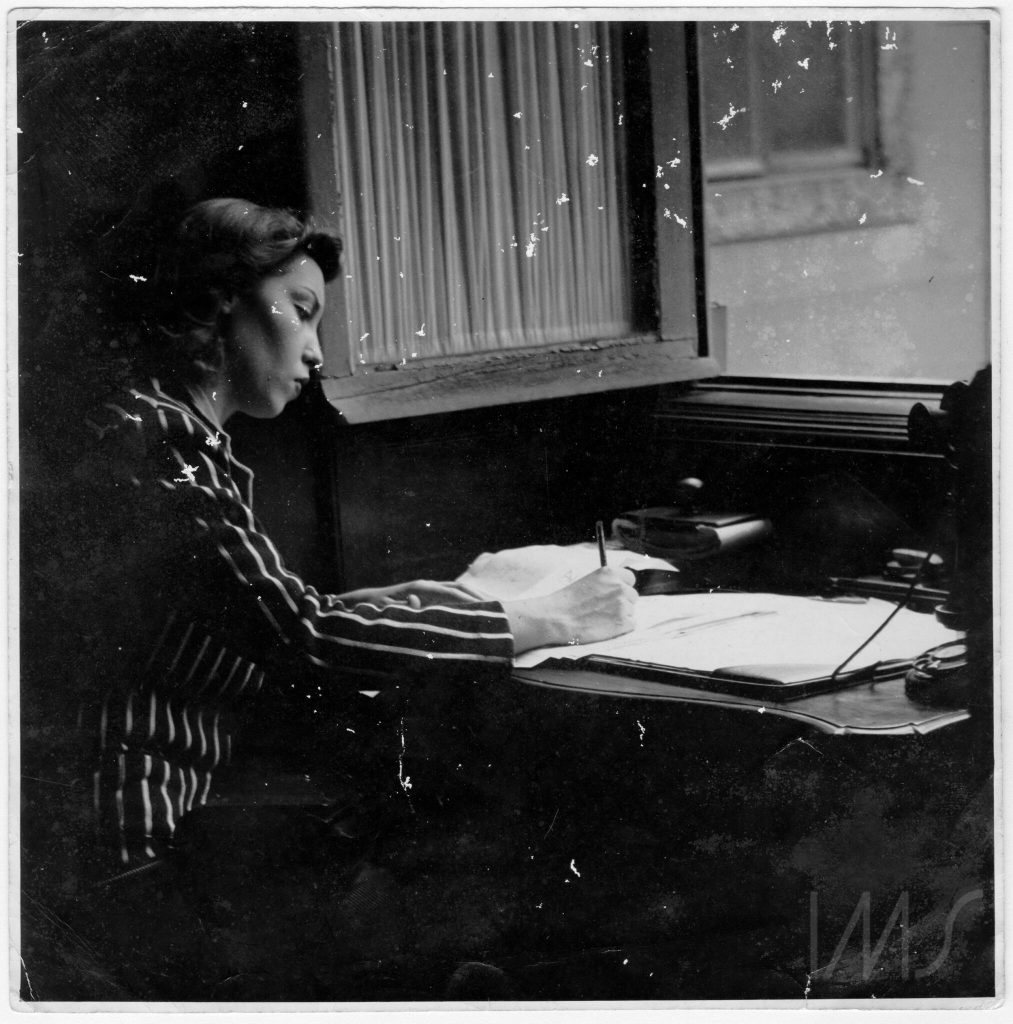 Black and white photograph of Clarice Lispector writing at her desk wearing a striped suit