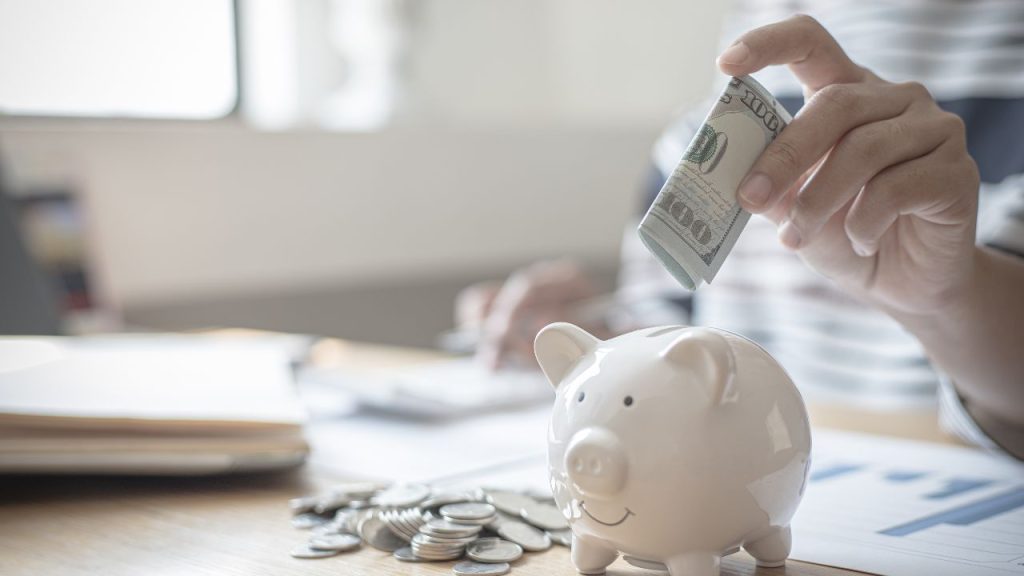 A hand putting a 100 dollar bill into a piggy bank.