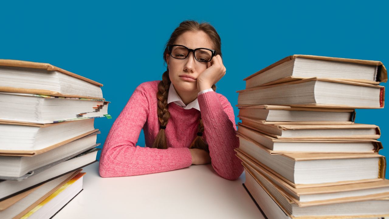A girl in glasses staring ahead in disappointment with stacks of books on either side of her.