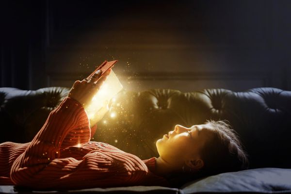 Girl laying face up on a couch reading from a glowing book.