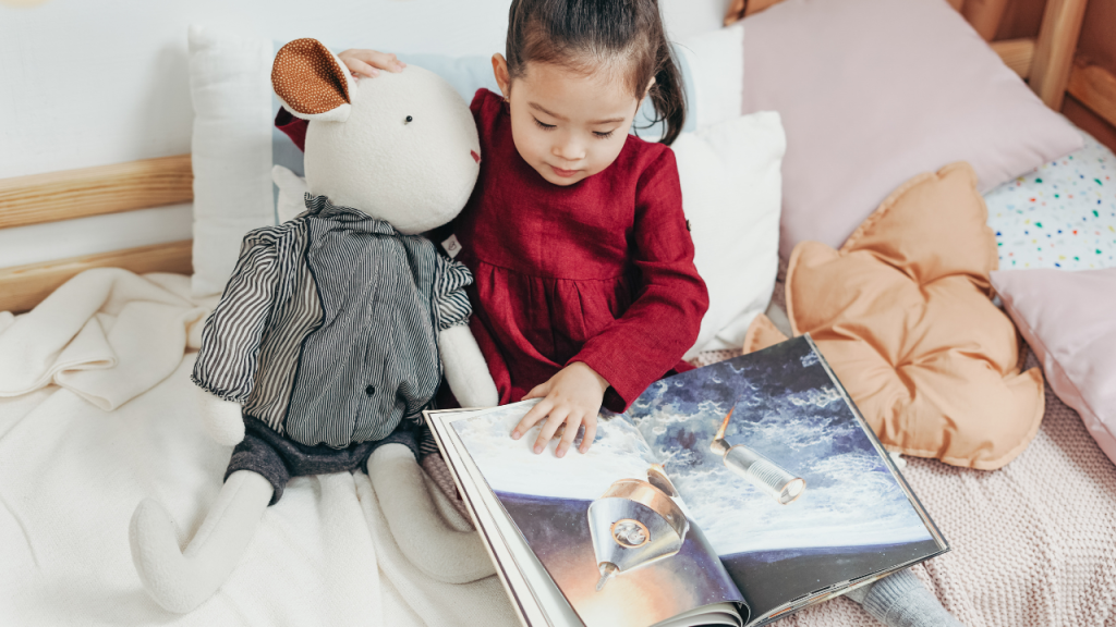 Child with book and stuffed animal on bed.