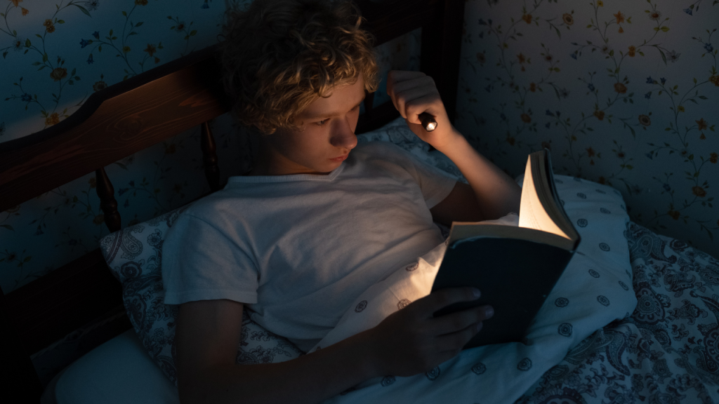Shows a person reading alone in the dark with flashlight.