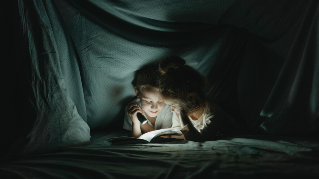 Children reading under bed sheets with flashlight.