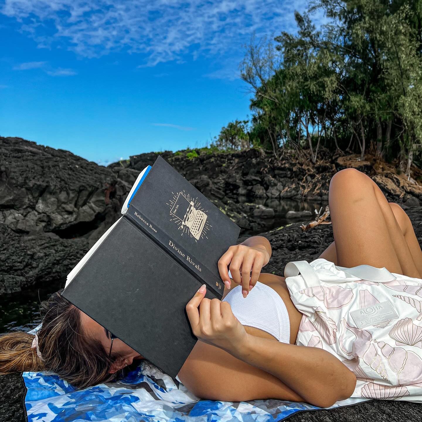 Leira Joyce laying on rocks reading a copy of Divine Rivals