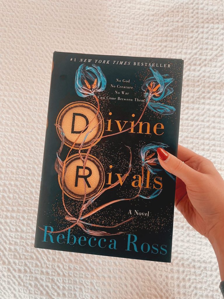 Sarah's right hand holding a copy of Divine Rivals over a white blanket