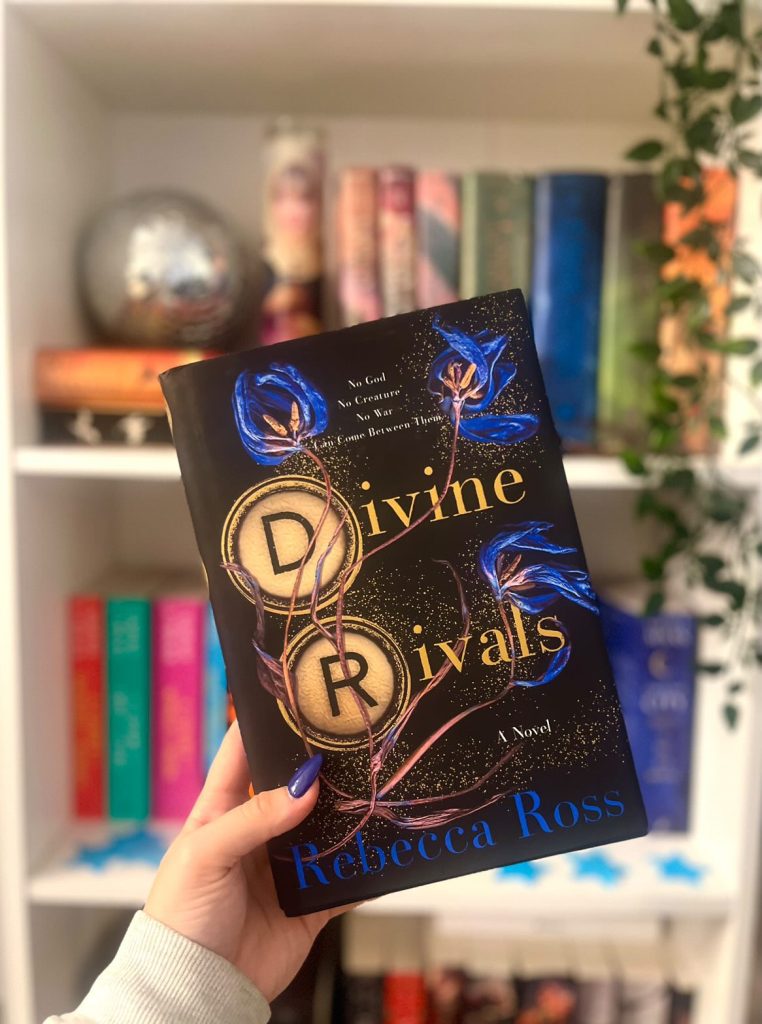 Keana's left hand holding a copy of Divine Rivals in front of her bookshelf