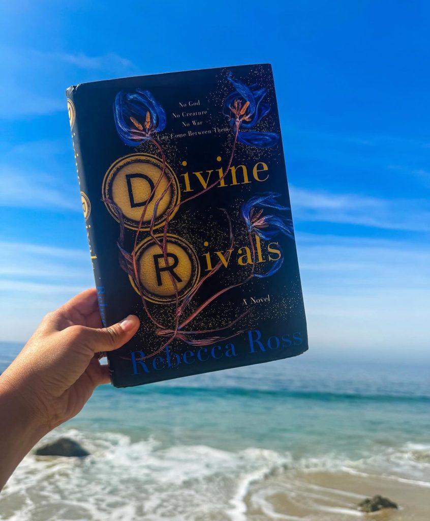 Karen holds Divine Rivals book against a backdrop of sunny beach and ocean.