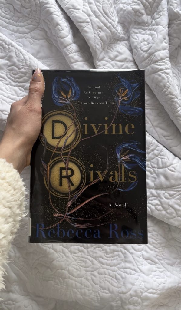 Steph D.'s left hand holding a copy of Divine Rivals over a white blanket