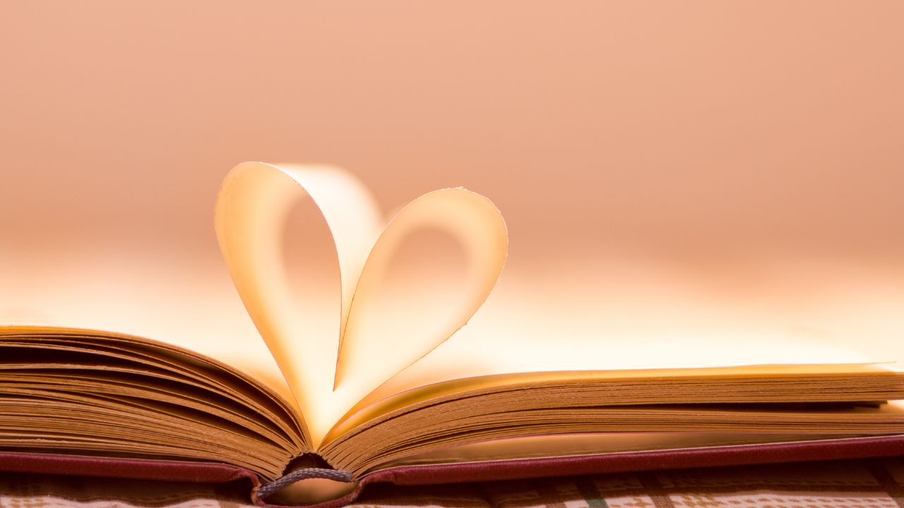 An open book lies slightly to the left as pages are folded into the shape of a heart. Set against a pinkish-orange background.