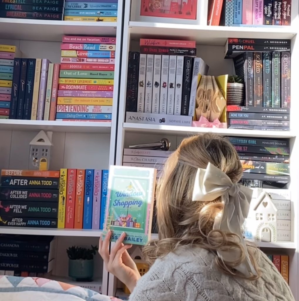 Hattie holding a copy of Window Shopping by Tessa Bailey while looking at her bookshelves