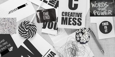 A black and white image of words, scribbles on paper, a striped pen, and a swirling lollipop sit against a gray surface. The words are splayed across the foreground in a chaotic fashion.