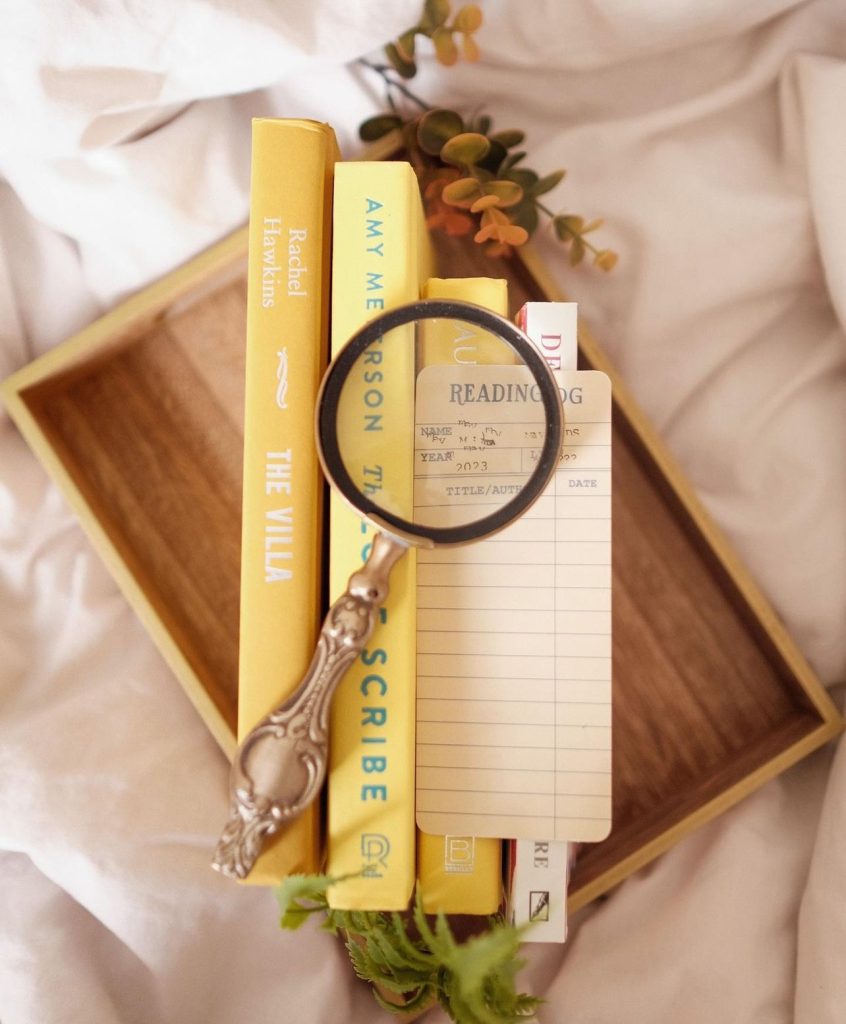 A magnifying class set on top of the spines of 4 yellow themed books that are piled on a wooden tray