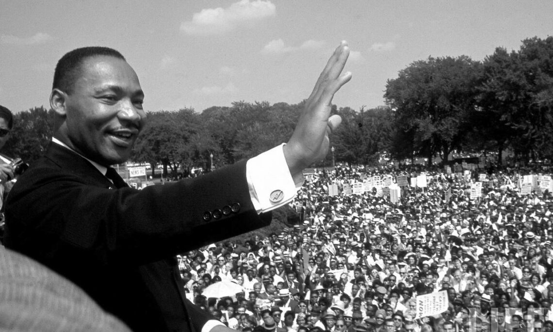 Black and white of Martin Luther King Jr. waving to a crowd of people. He and the crowd are outside. There are trees behind them. They seem to be a park.