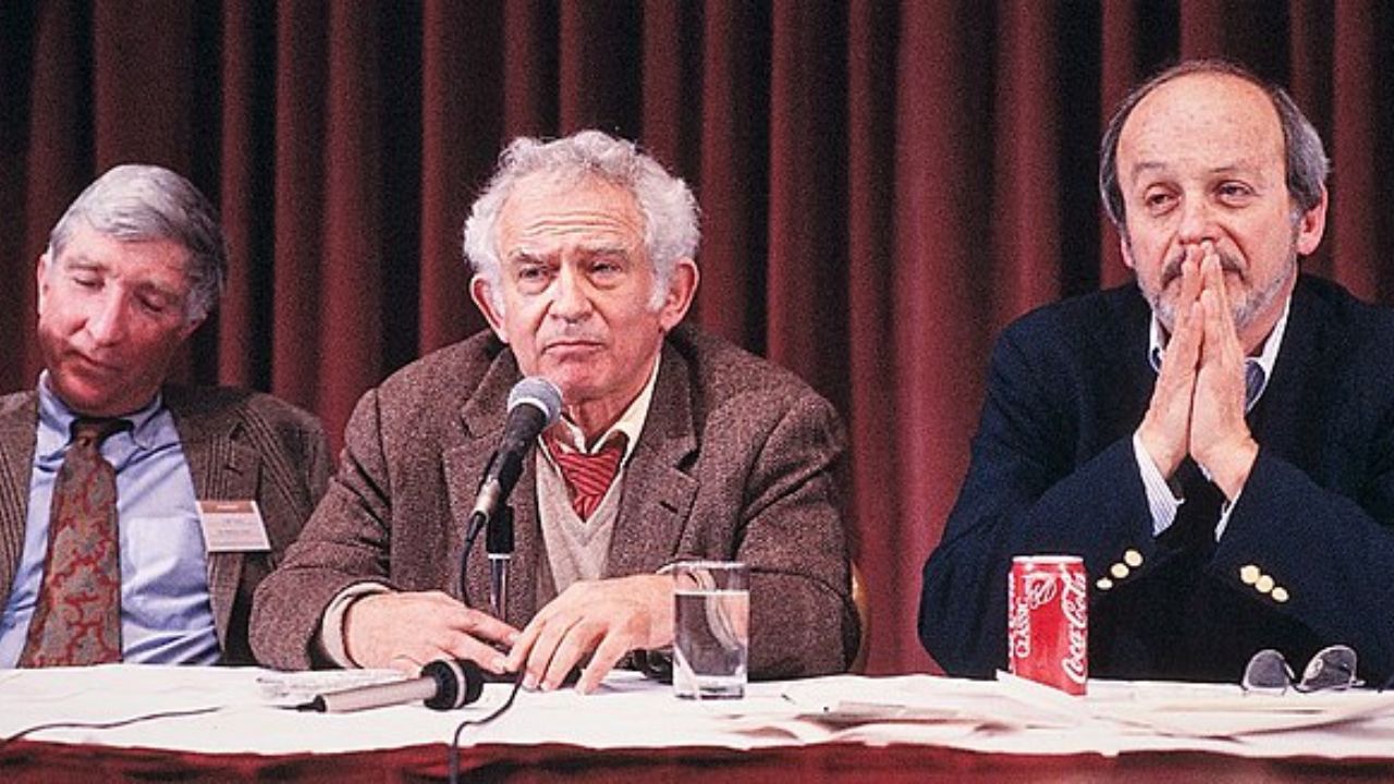 At a PEN conference, Norman Mailer sits between two men all in suits, in front of a red curtain. A microphone, scattered papers, and a soda sit on the table in front of them.