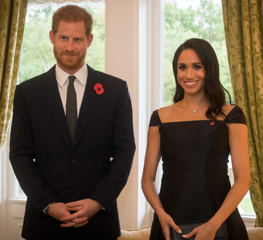 Image showing Prince Harry dressed in suit and tie with red flower pin on his lapel. He is standing indoors next to his wife, Meghan Markle, who is dressed in a formal black dress with matching red flower pin. 