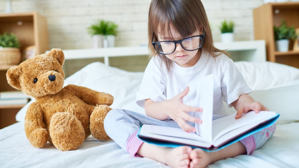 Image of a child and a stuffed bear reading a book on a bed. 