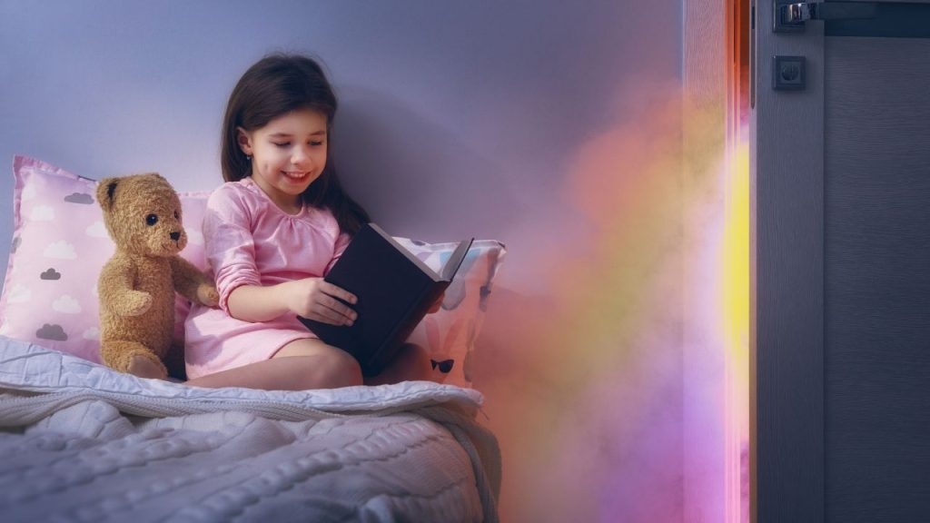 Image of a child and bear in bed reading a book with a door spilling out a rainbow and a magical beam. 